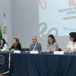 Mesa com toalha azul, tendo uma tela projetada na parede ao fundo com 10º Encontro Nacional do Cogecom. Sentados à mesa, estavam cinco pessoas que falaram na abertura, quatro mulheres e um homem.