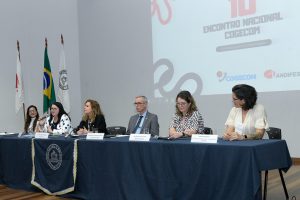 Mesa com toalha azul, tendo uma tela projetada na parede ao fundo com 10º Encontro Nacional do Cogecom. Sentados à mesa, estavam cinco pessoas que falaram na abertura, quatro mulheres e um homem.